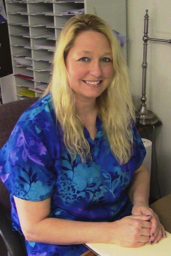 A woman with blonde hair is smiling at her desk in an office setting, wearing a blue shirt.