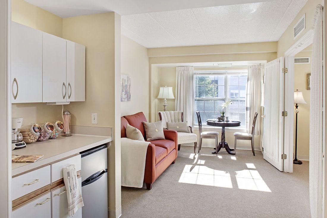 A cozy apartment living space with a kitchenette, sofa, and small dining area by a sunlit window.