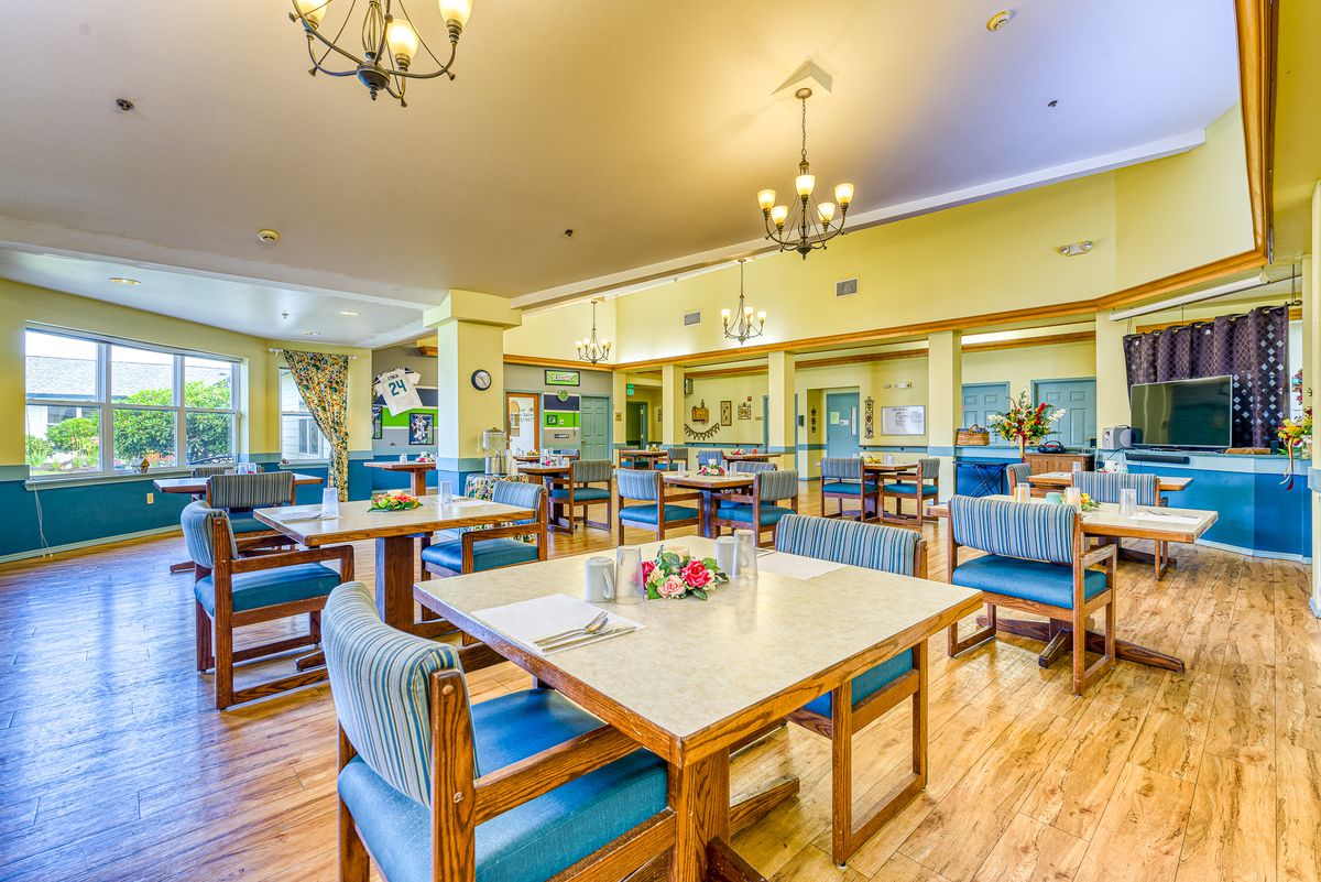 A brightly lit dining area with neatly arranged tables and chairs, featuring elegant chandeliers and wooden flooring.