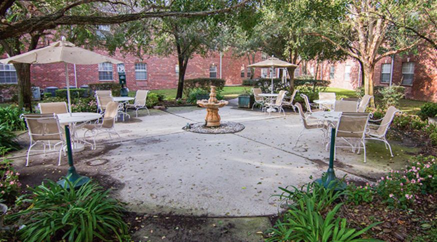 A serene outdoor patio area featuring a central fountain, surrounded by tables and chairs under shady trees, with a brick building in the background.