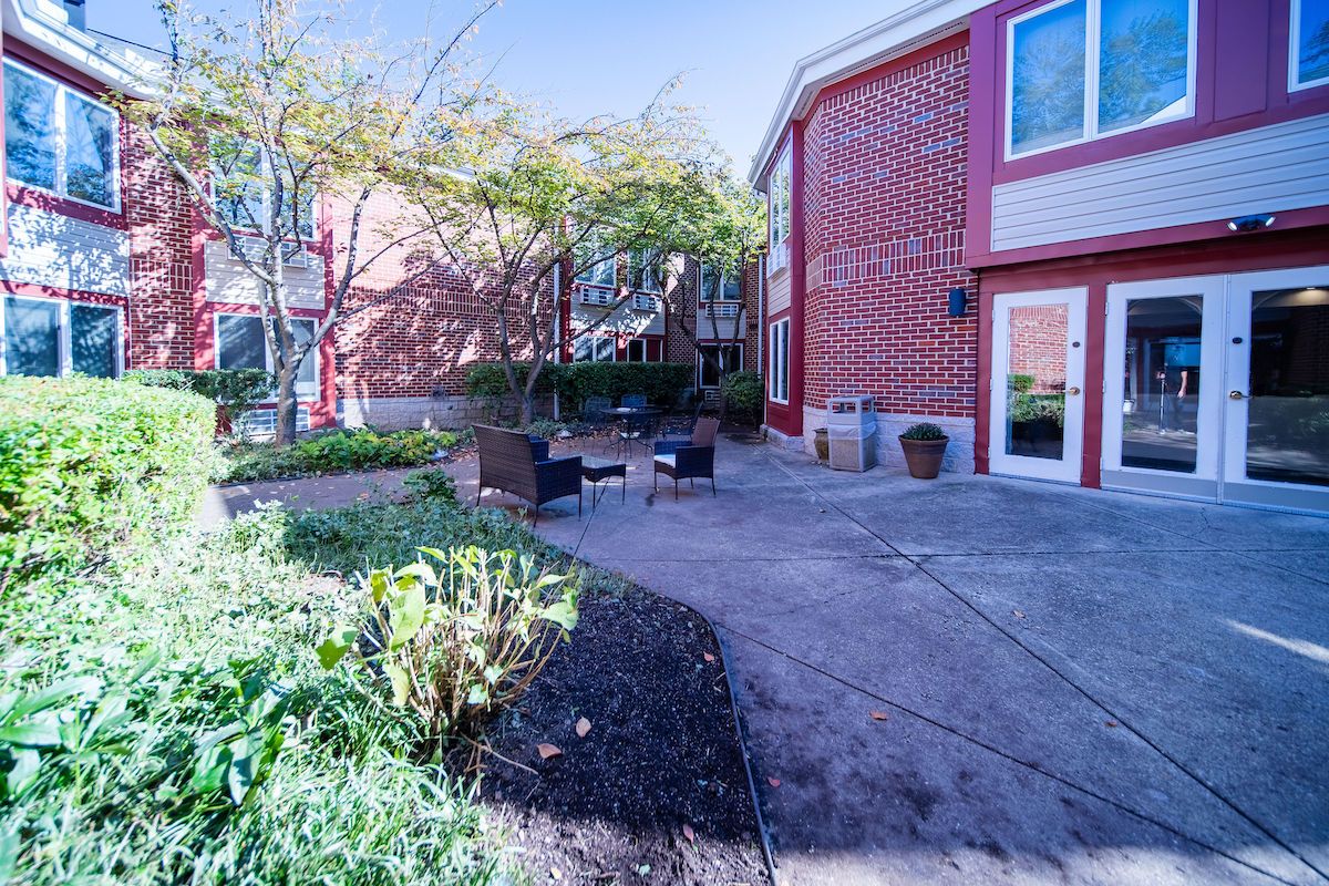 A charming outdoor patio area with seating, surrounded by greenery and adjacent to a red-brick building.