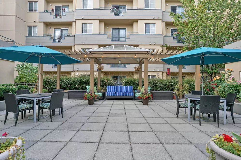 A courtyard with outdoor seating, featuring a pergola and umbrellas, in front of an apartment building.