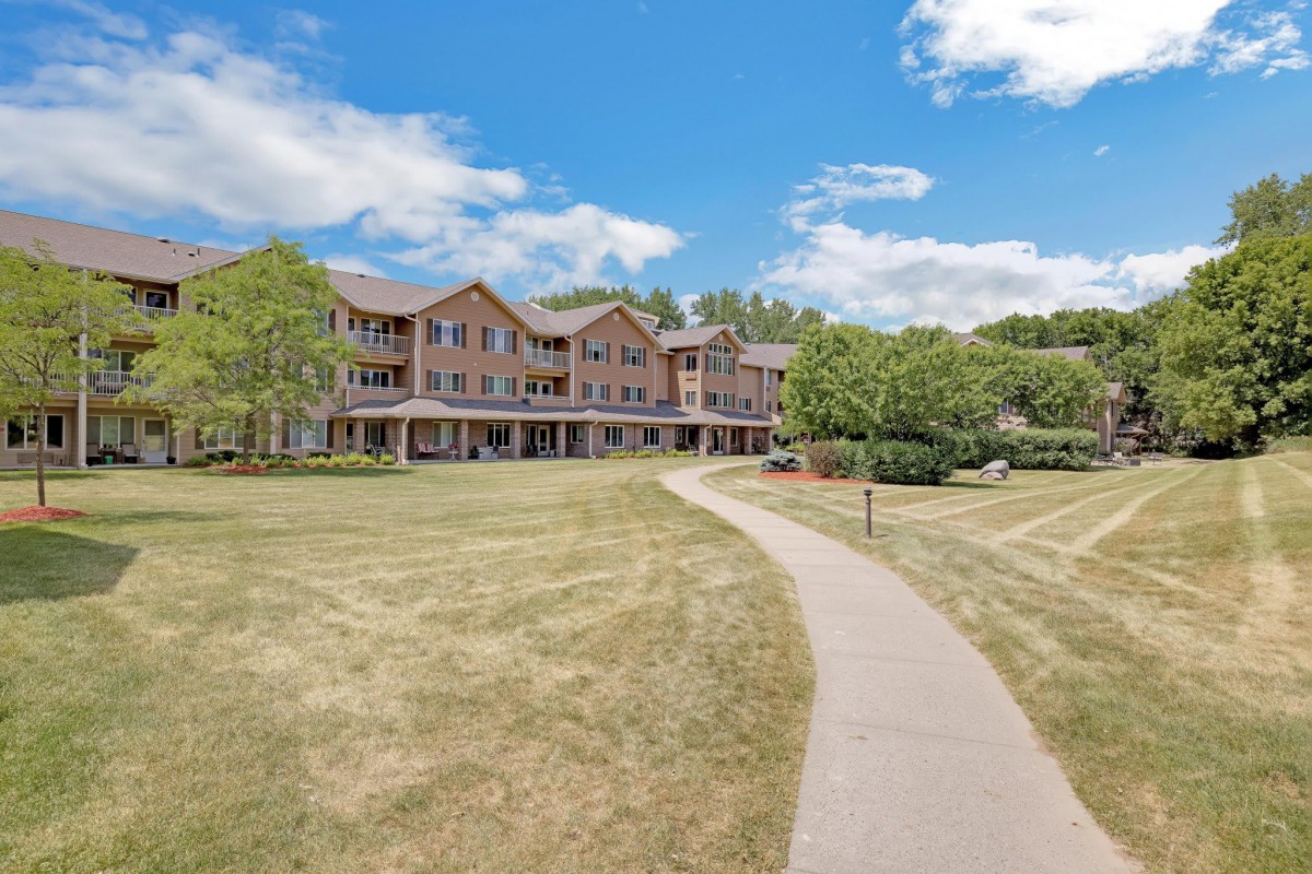 A scenic view of a large building with a pathway leading through a grassy lawn surrounded by trees under a blue sky with clouds.