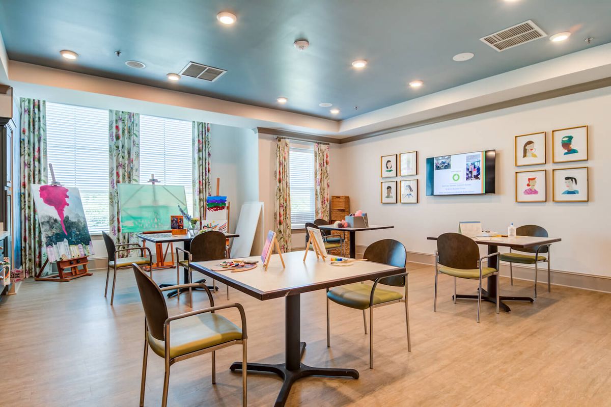 A modern, well-lit room with tables, chairs, and art supplies near large windows.