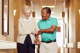 A caregiver assisting an elderly person as they walk together indoors, both smiling.