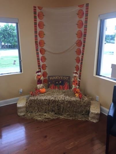 An indoor fall-themed display featuring hay bales, scarecrows, and pumpkins, framed by festive autumnal decorations.