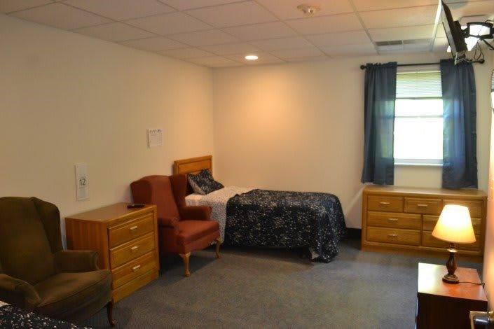 A simple bedroom with a single bed, armchairs, and wooden dressers.