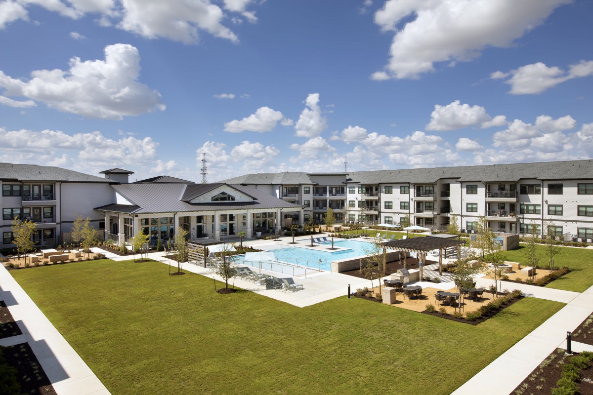A modern apartment complex with a central swimming pool and a well-maintained lawn under a partly cloudy sky.