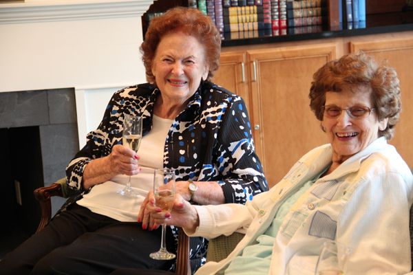Two elderly women smiling and holding hands while enjoying drinks indoors.