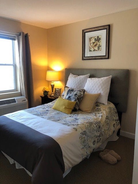A cozy bedroom with a neatly made bed, decorative pillows, a bedside lamp, and a window providing natural light.