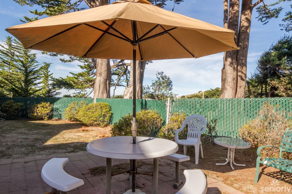 A cozy outdoor patio area with a round table, umbrella, and chairs surrounded by greenery.