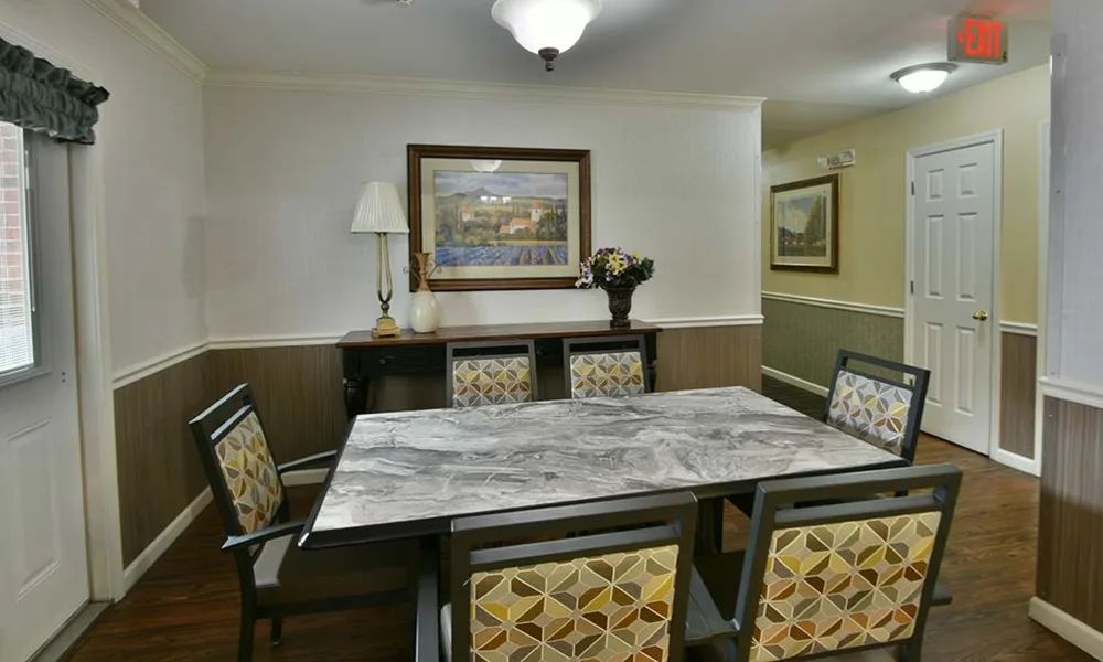 A cozy dining room with a marble-topped table and patterned chairs, adorned with a painting, lamp, and vase.
