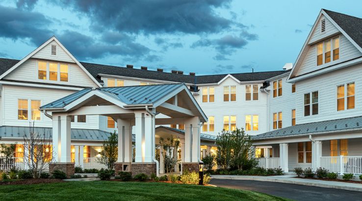 A large residential building with a well-lit entrance and landscaped surroundings under a cloudy evening sky.
