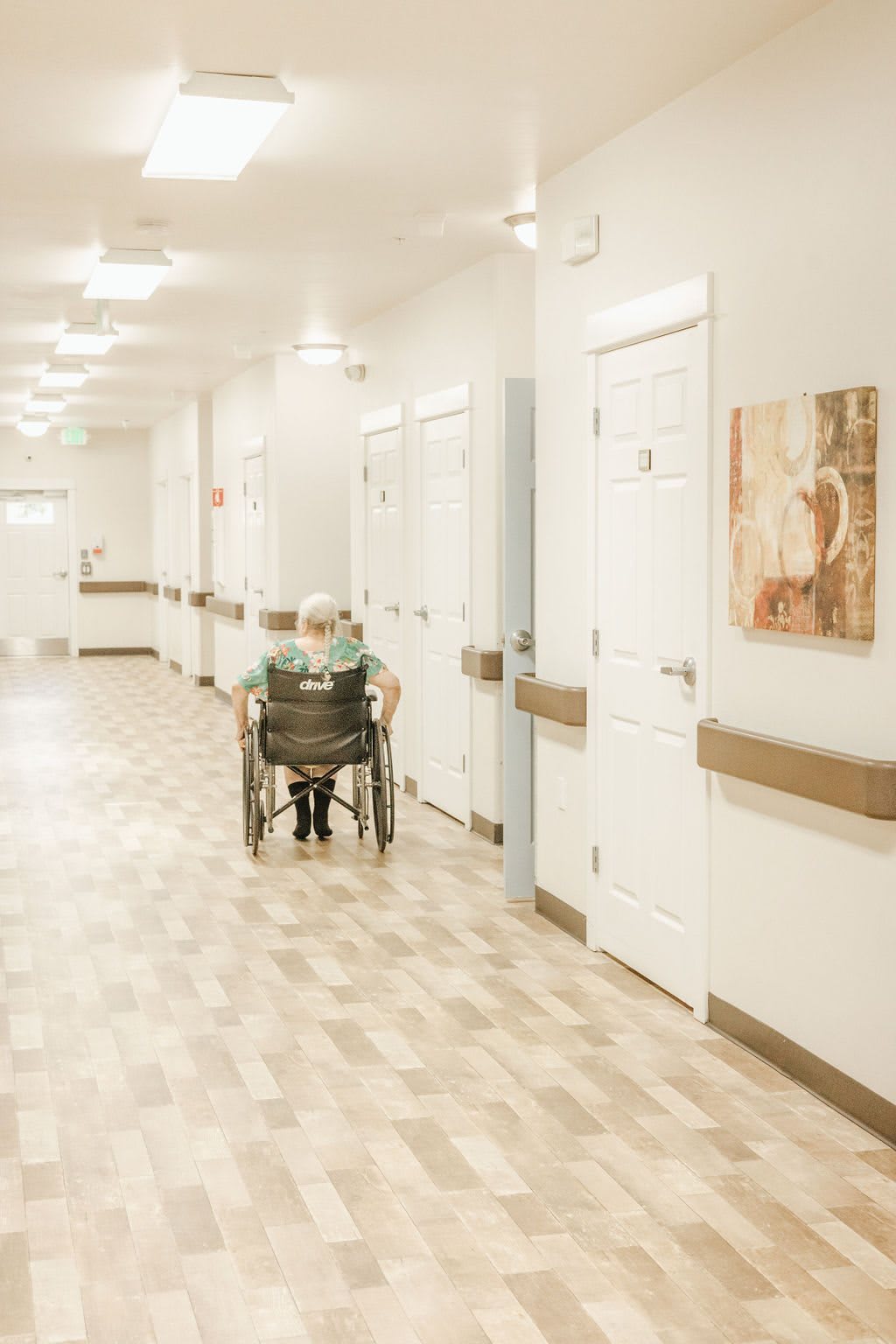 An elderly person in a wheelchair sits in a long hallway of what appears to be a nursing home.