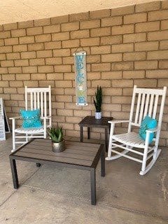 A cozy patio setup with white rocking chairs, decorative pillows, and plants against a brick wall.