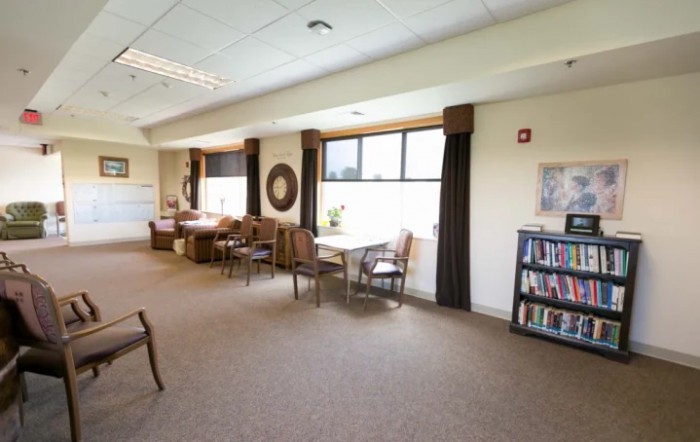 A spacious lounge area with seating and a bookshelf, illuminated by natural light through large windows.