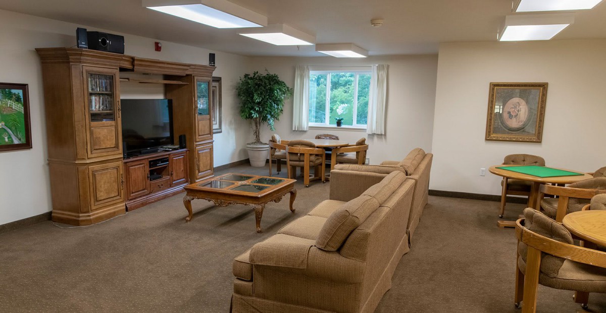 A cozy living room featuring a sofa, coffee table, and entertainment center with a television. A dining table is set near the window, complemented by an indoor plant.
