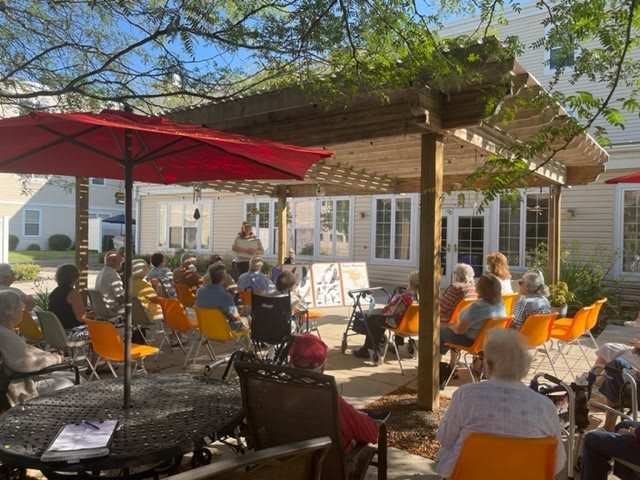 A sunny outdoor event with an audience seated under a canopy.