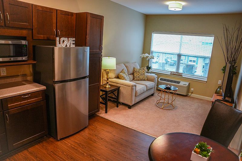 A cozy apartment living room with a kitchen area, featuring a comfortable sofa, large window, and wooden flooring.