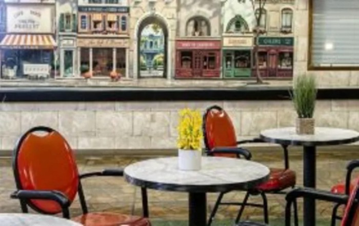 An outdoor cafe with empty tables and chairs, adorned with a pot of yellow flowers, against a backdrop of charming street facades.