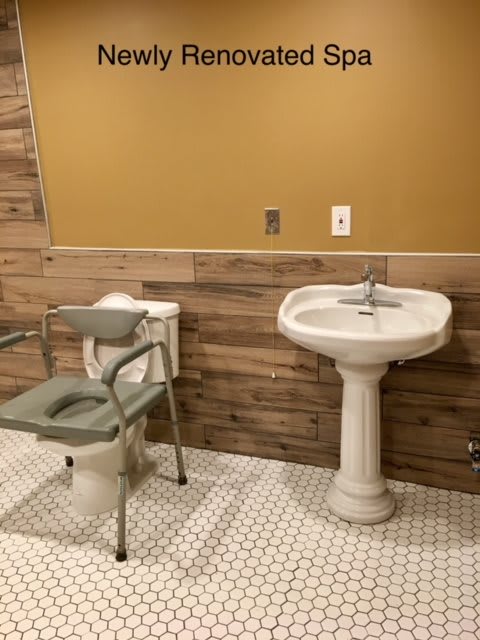 A bathroom featuring a pedestal sink and a toilet with a commode chair, set against wood-paneled walls and tile flooring.