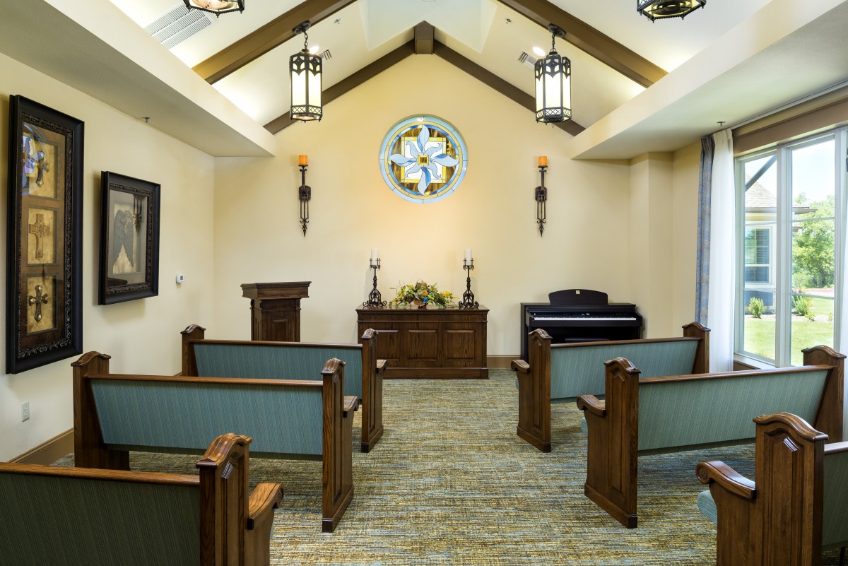 A peaceful chapel interior featuring wooden pews, an altar with candles and flowers, a stained glass window, and a piano.