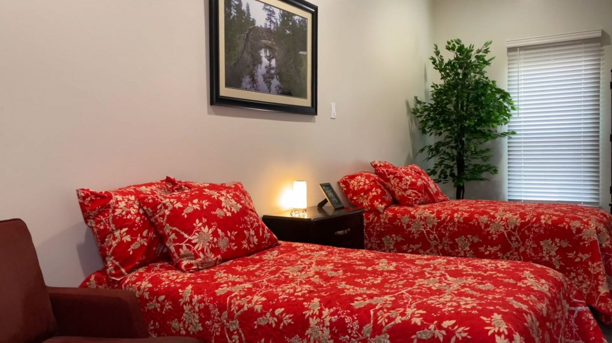 A cozy bedroom with red-patterned bedding, a nightstand with a small lamp, and decorative artwork on the wall.