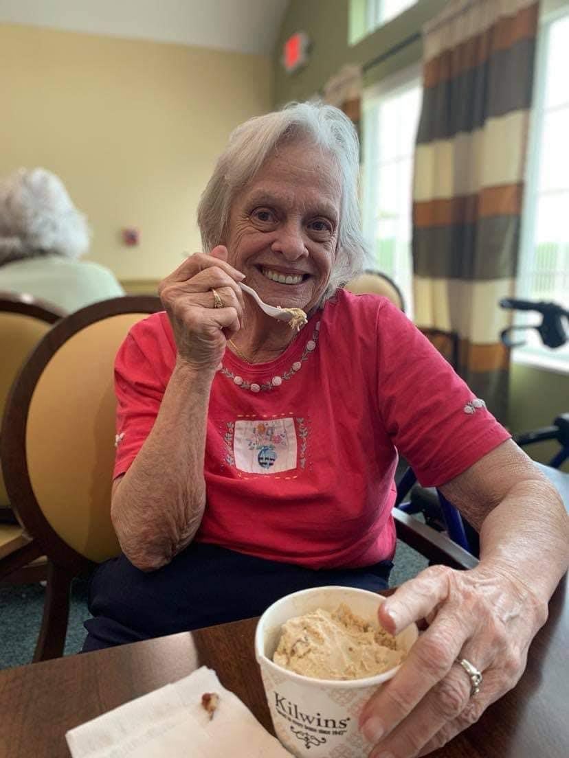 An elderly woman enjoys ice cream while smiling indoors.
