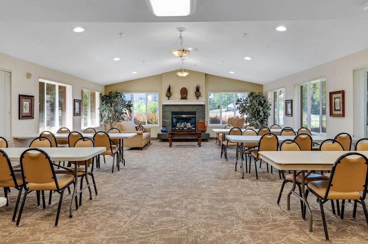 A spacious community center meeting room with tables and chairs, featuring a fireplace and large windows.