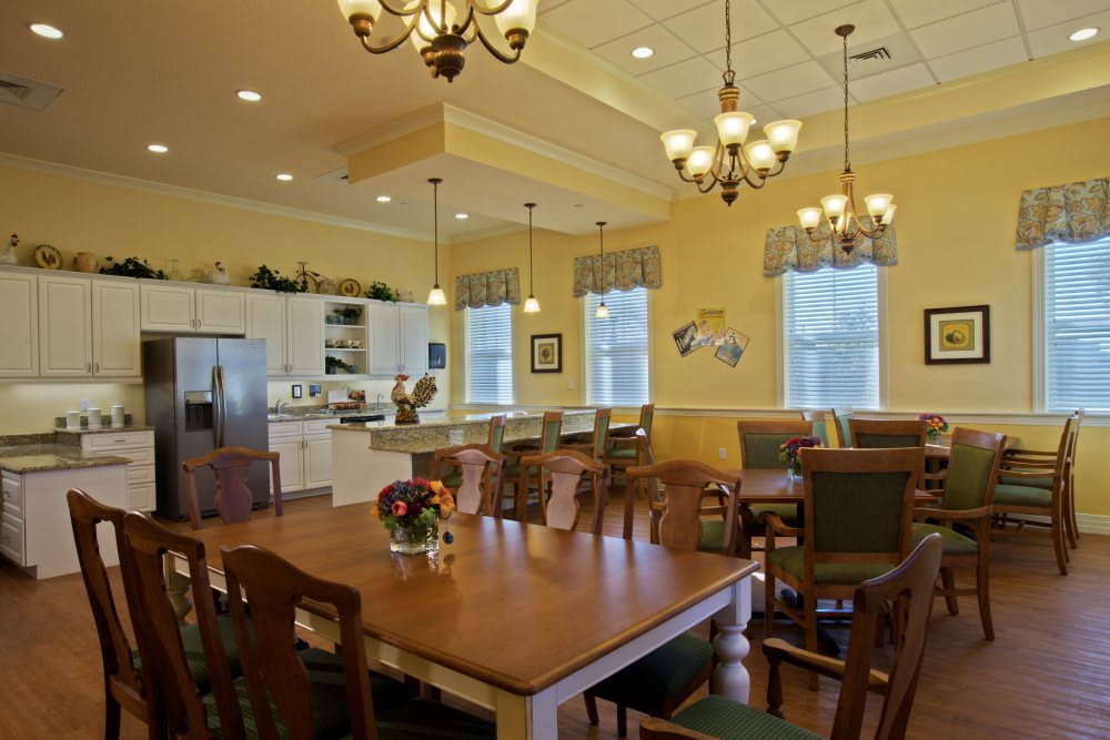 A spacious dining area with a wooden table and a kitchen in the background, featuring elegant chandeliers and large windows.