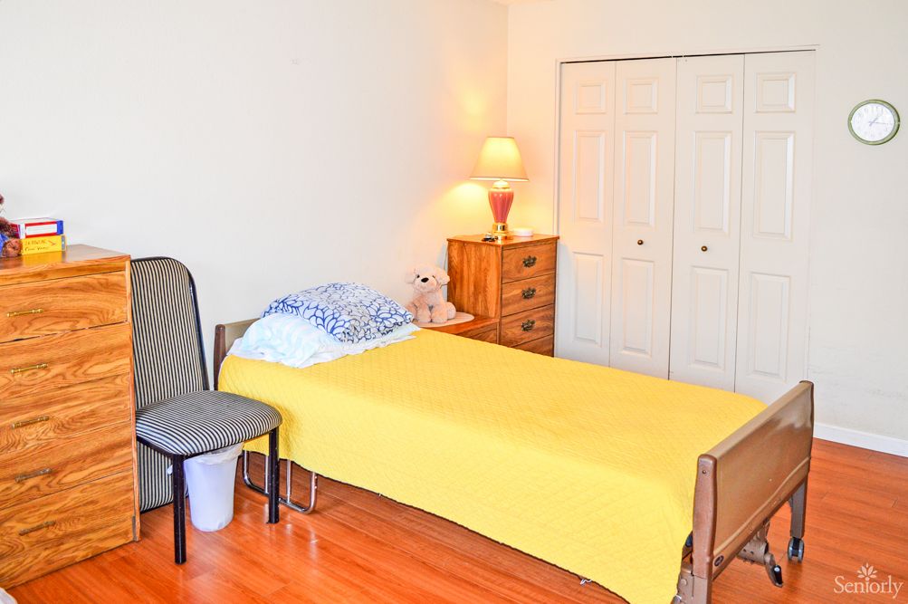A simple bedroom with a single bed covered in a yellow blanket, a wooden dresser, and a lamp on top.