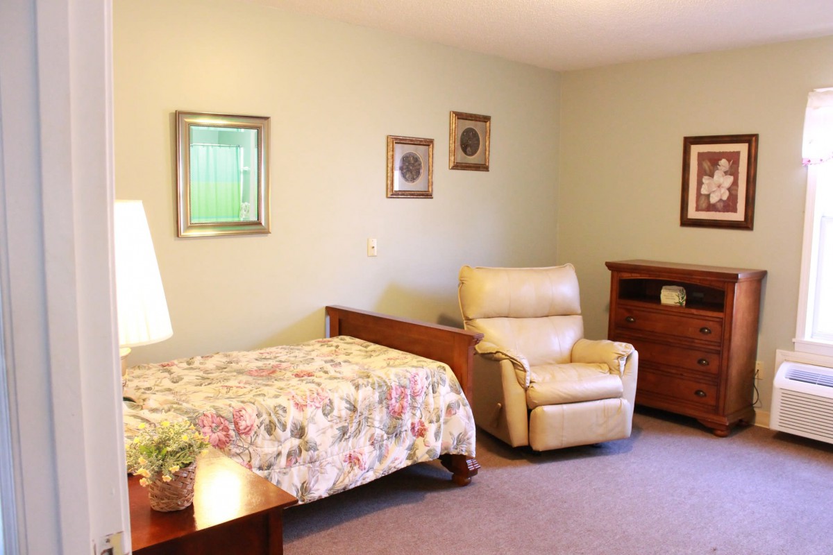 A cozy bedroom featuring a bed with floral bedding, an armchair, framed wall art, and a lamp.