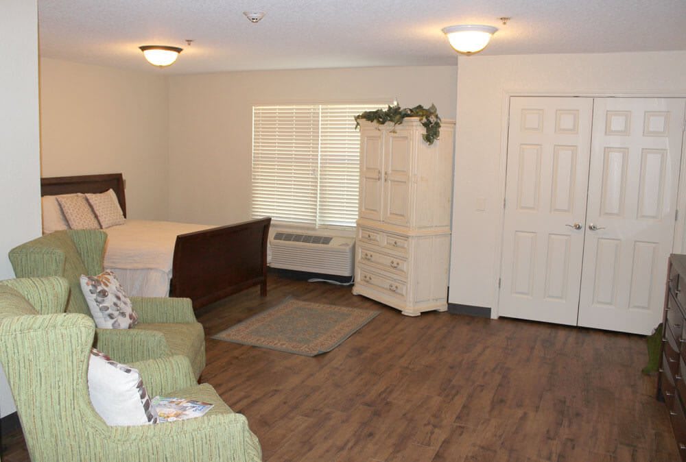 A cozy bedroom with a wooden floor, featuring a bed, dresser, and armchairs.