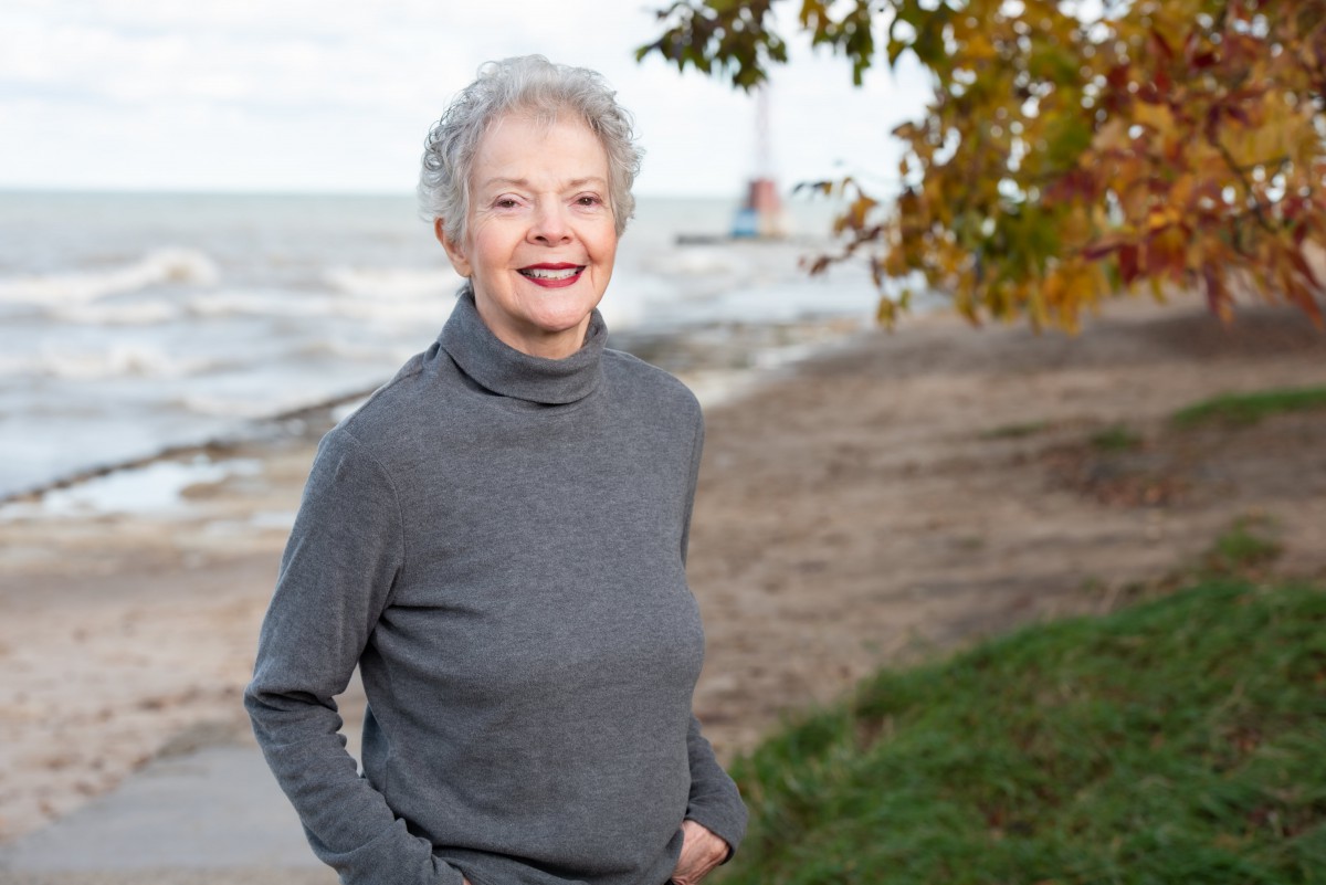 A woman in a grey sweater stands smiling by the beach in autumn.