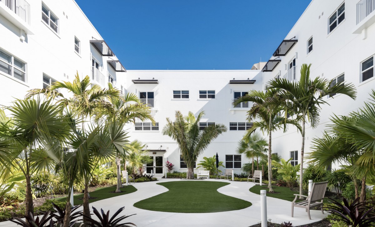 A modern white building with a landscaped courtyard featuring palm trees under a clear blue sky.