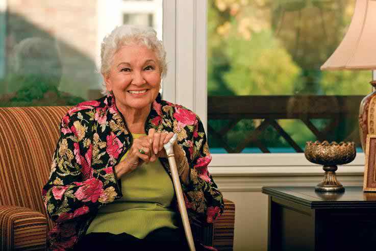 An elderly woman with a bright smile sits indoors, holding a cane and wearing a floral jacket.