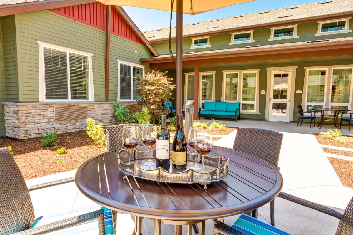 A sunny patio with a round table set for wine tasting, featuring three glasses and two bottles of red wine under an umbrella in a cozy backyard setting.