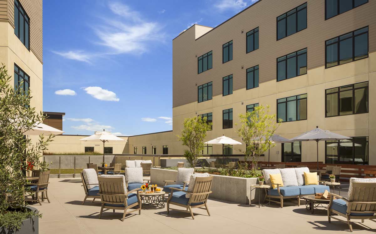 A modern outdoor patio area with comfortable seating and umbrellas, set against a multi-story building under a clear blue sky.