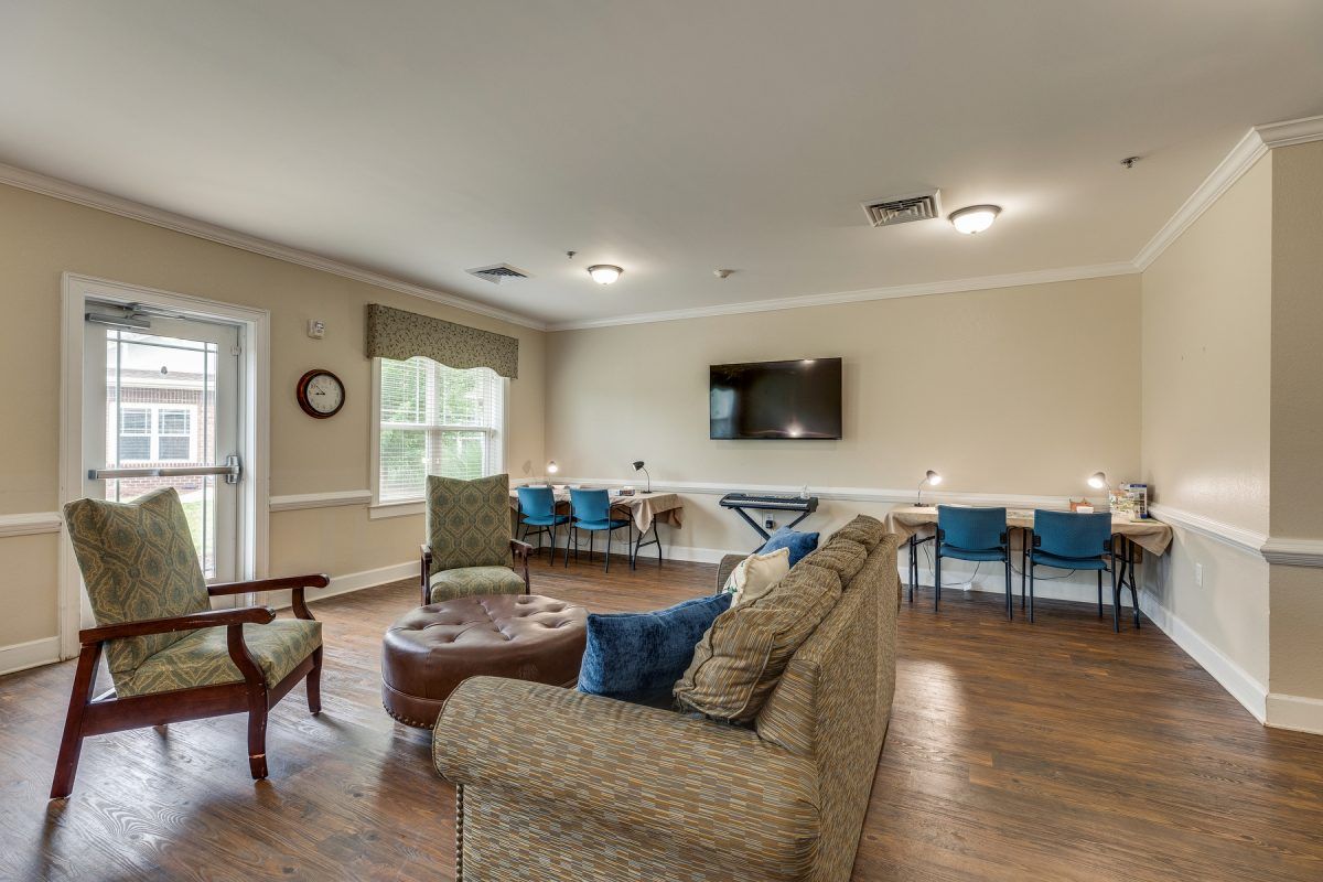 A spacious living room featuring comfortable seating, a TV on the wall, and a row of chairs along the back wall.
