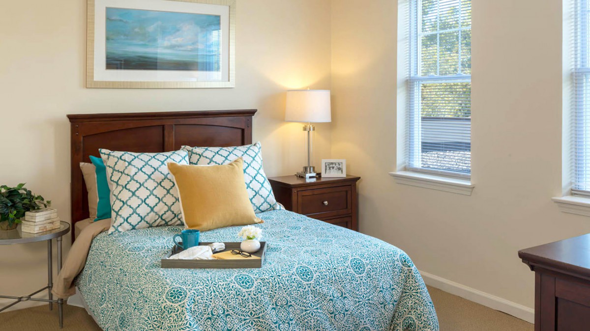 A cozy bedroom with a neatly made bed, featuring patterned bedding and a decorative pillow. A lamp sits on the nightstand next to the window.