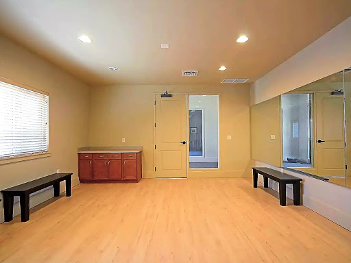 A spacious, empty room featuring wooden flooring, benches, and natural light pouring in from the windows.