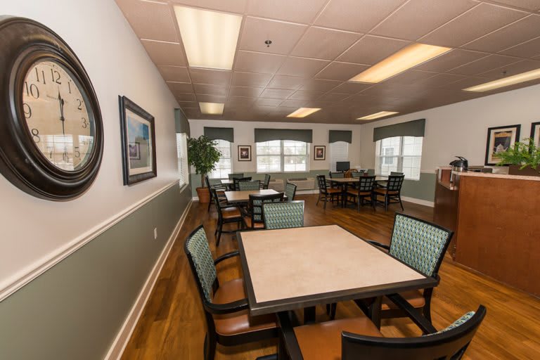 A well-lit dining area with multiple tables and chairs arranged neatly, featuring large windows and a wooden floor.