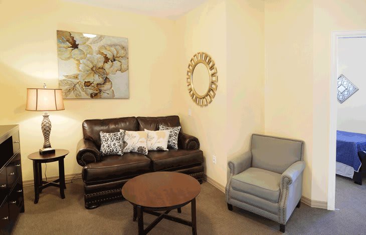 A cozy living room with a brown leather sofa, a light blue armchair, and a round coffee table. The walls are adorned with floral artwork and a decorative mirror.