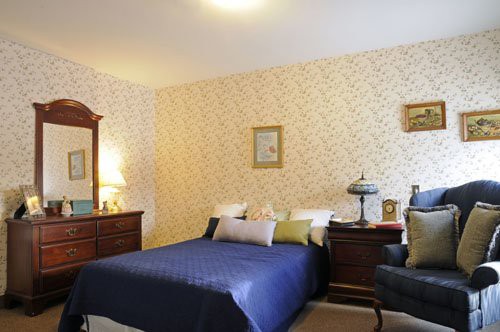 A cozy bedroom with a classic design, featuring a blue bedspread, wooden furniture, and decorative wallpaper.