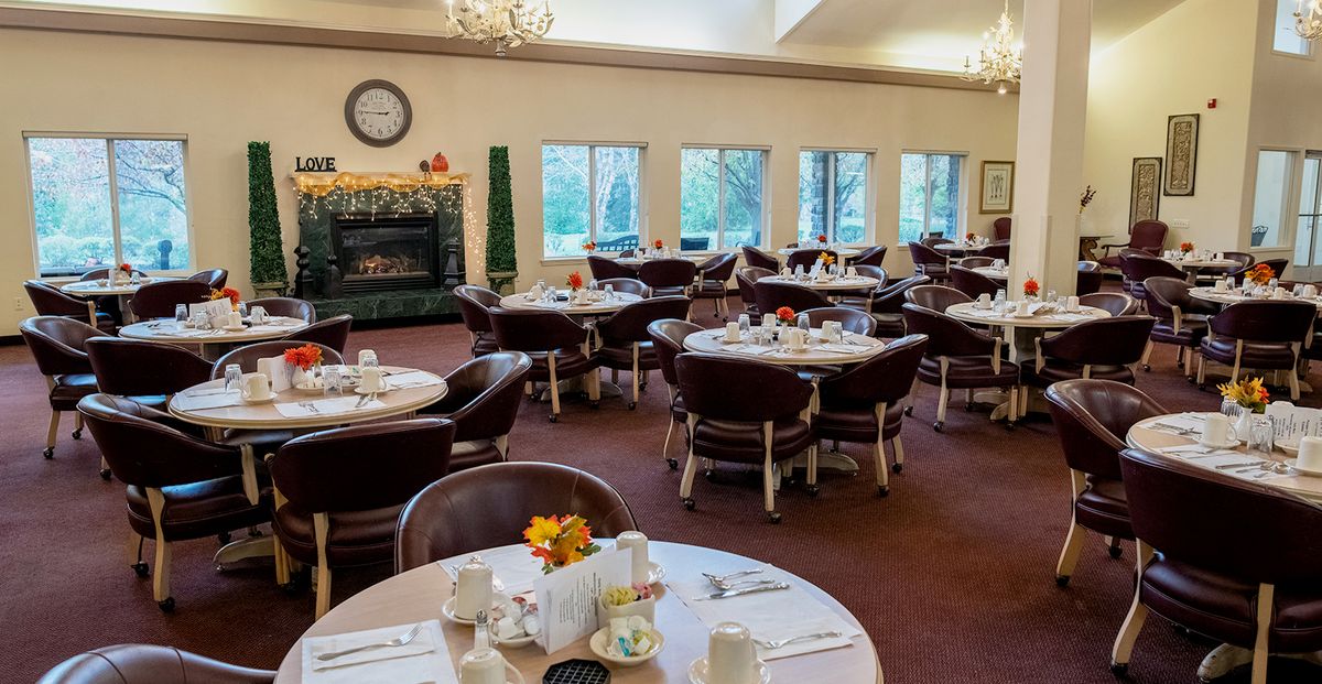 A cozy dining room with neatly set tables and chairs, featuring a fireplace and large windows.