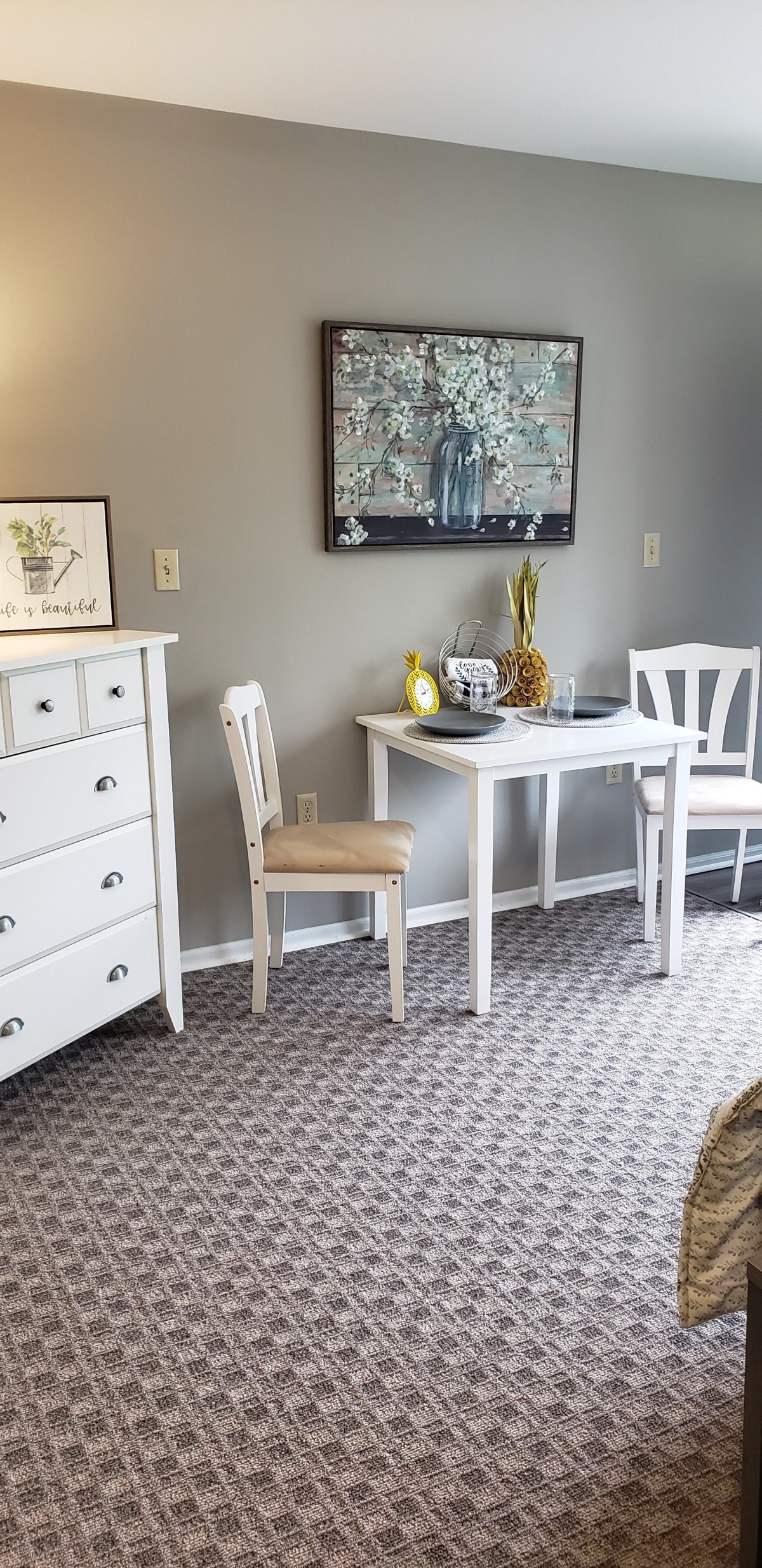 A cozy dining area with white furniture, including a table and chairs, next to a dresser adorned with decor and artwork.