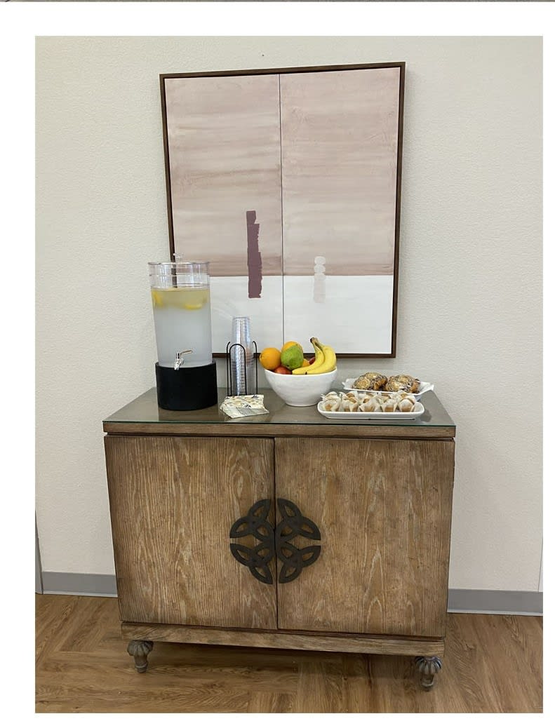A refreshment station featuring a beverage dispenser, fruit bowl, and cookies on a wooden cabinet with artwork above.