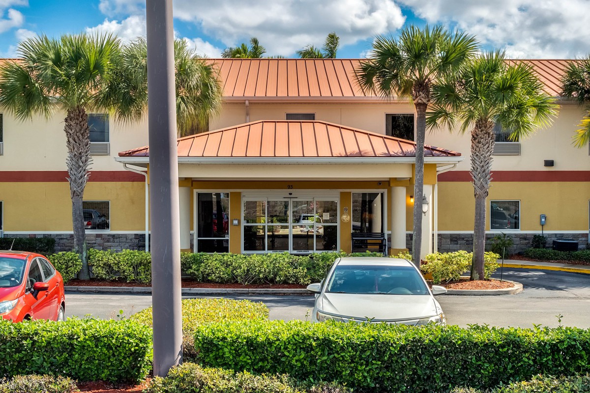 A building entrance with a car parked in front, surrounded by palm trees and landscaping under a partly cloudy sky.