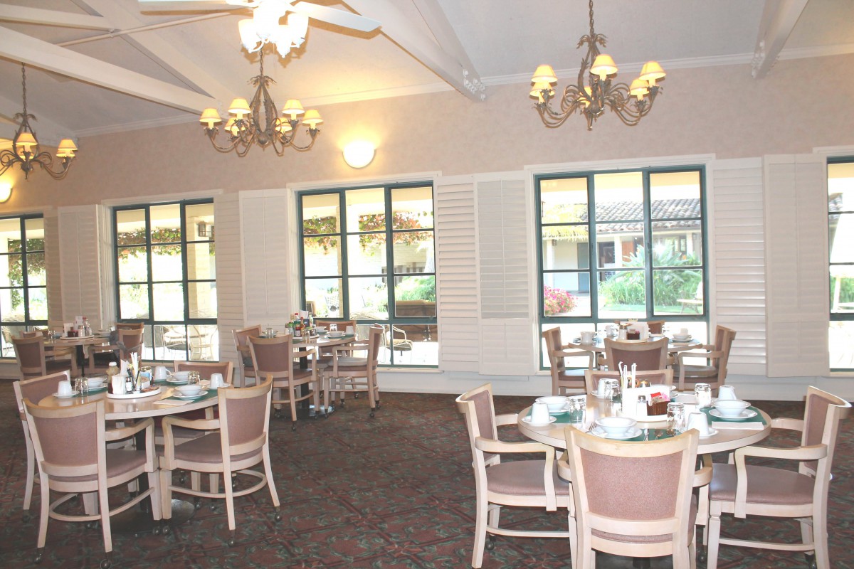 An empty dining room with chandeliers and neatly arranged tables and chairs near large windows.