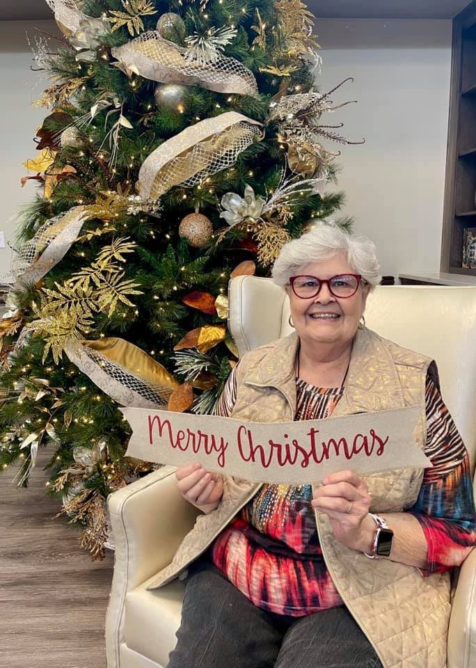 A woman sitting in front of a Christmas tree holding a 'Merry Christmas' sign.